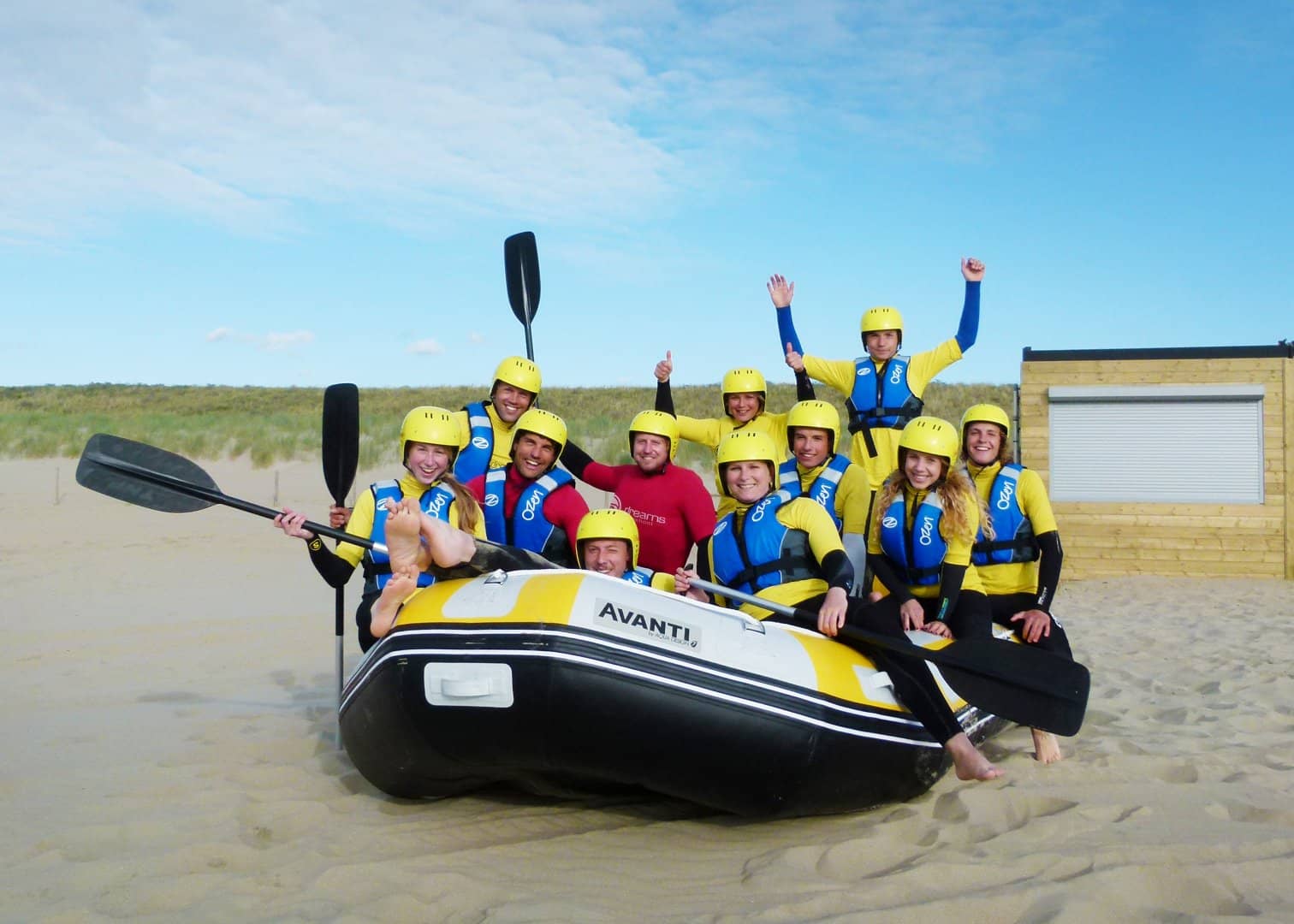 foto van een groep enthousiaste mensen die gaan raften in wetsuits en met gele helmen en roeispanen in een rubberen boot op het strand van Ter Heijde in het Westland