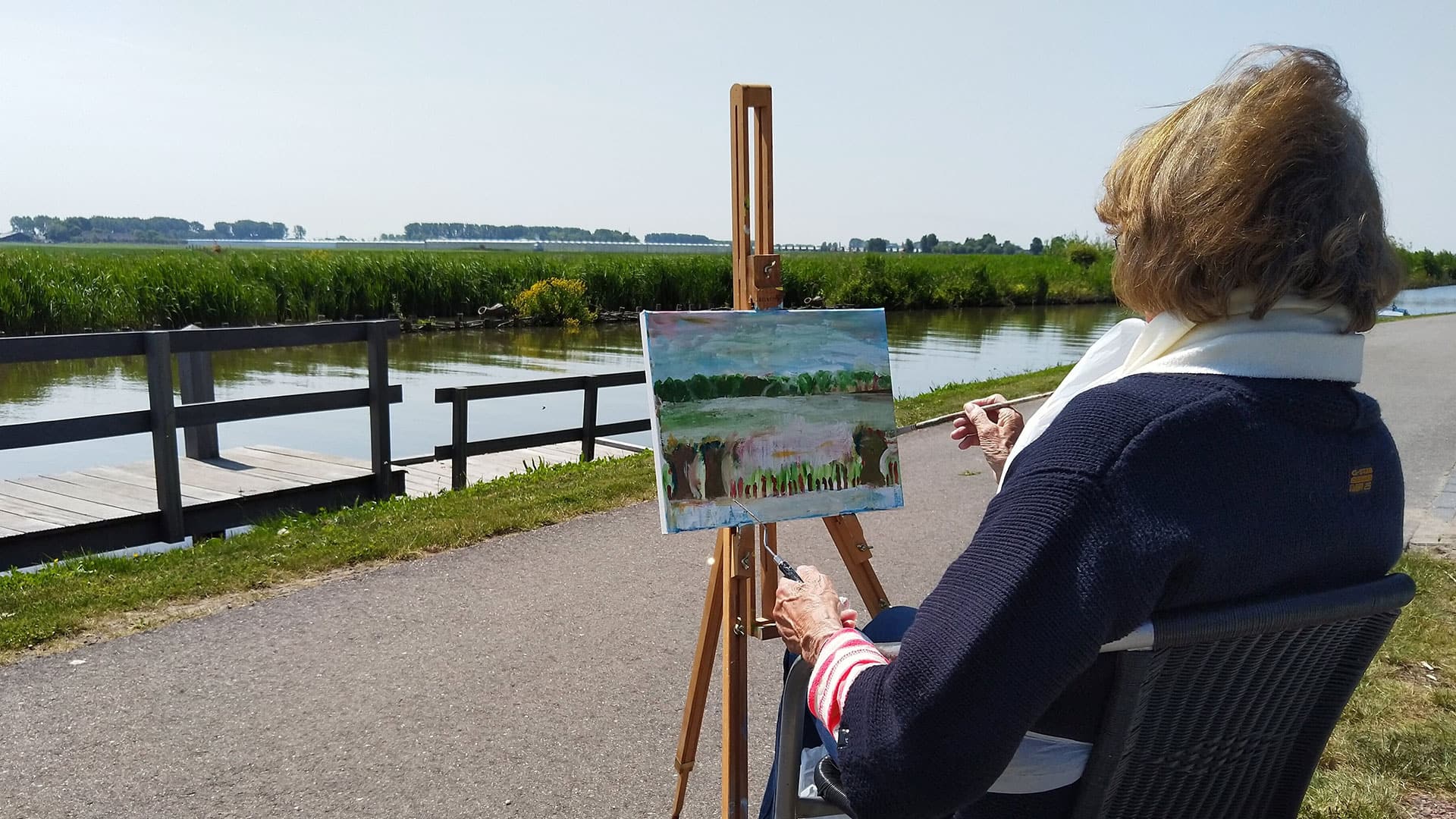 foto van een mevrouw en een schildersezel, op de rug bezien, die zit te schilderen langs de oever van de rivier de Zweth in het Westland