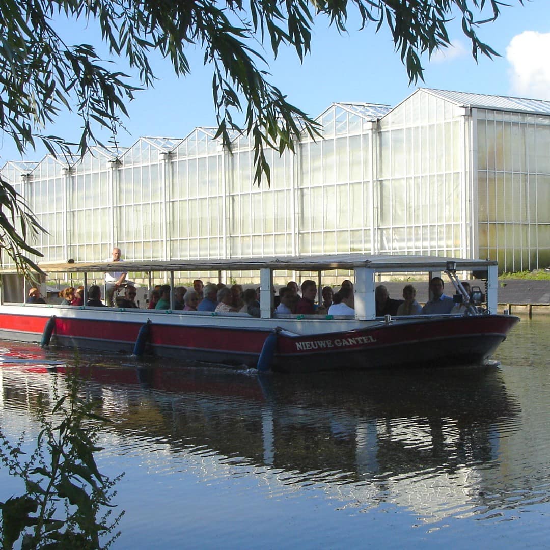 Rondvaartboot van Rondvaartbedrijf De Gantel vaart met gasten aan boord door het Westland met op de achtergrond glazen kassen in de zon