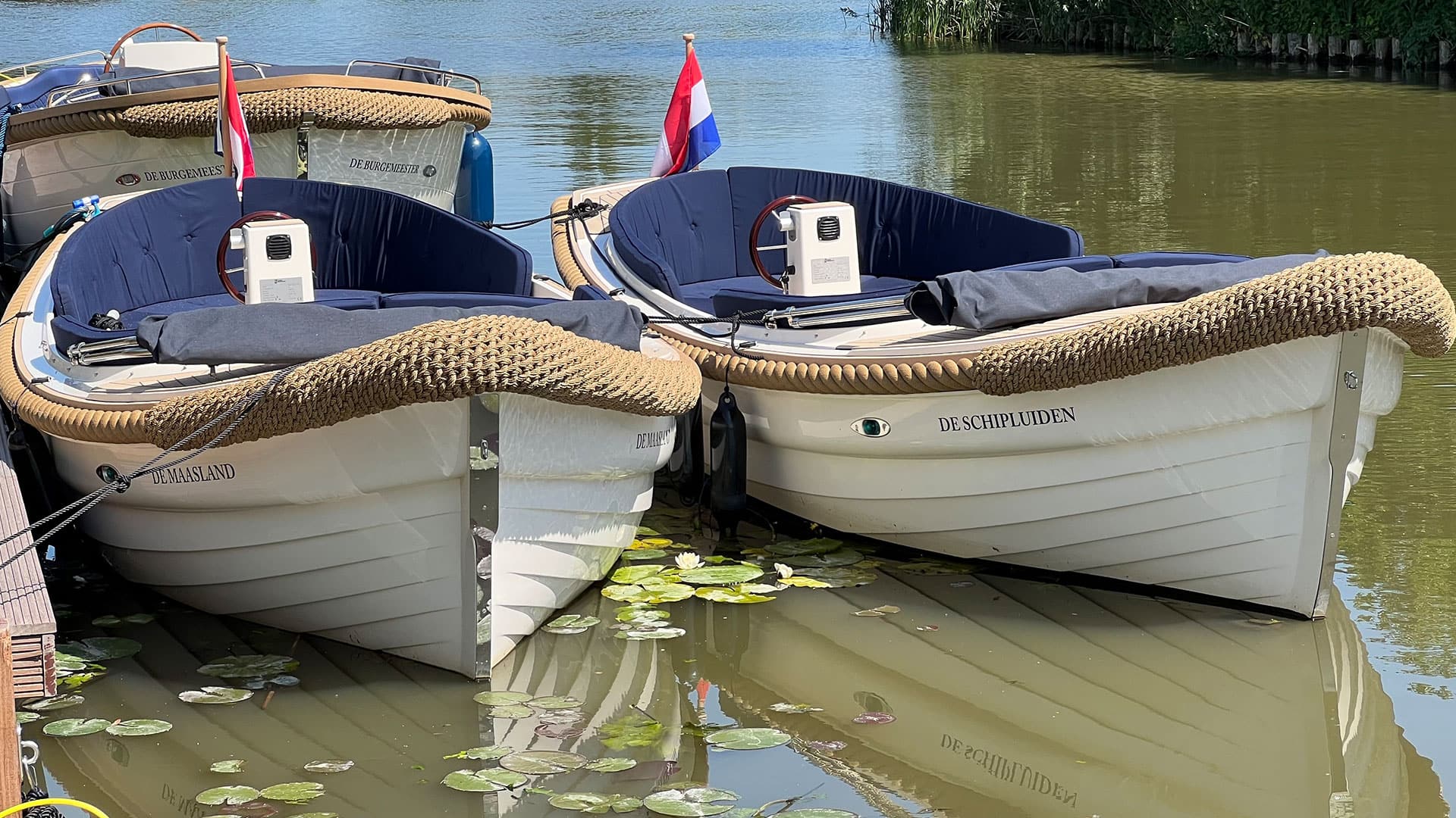 luxe elektrische sloepen van Floriette Experience en 1 motorsloep in het water met witte scheepsromp en blauwe kussens op een zonnige dag in Schipluiden voor een vaartocht naar het Westland