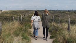 Twee vrouwen lopen naast elkaar door de duinen aan de Westlandse kust met in het midden een wandelpad en links en rechts met helmgras begroeide duinen