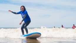 vrouw met lang donker haar in een zwart wetsuit en blauw t-shirt op een surfplank van Dutch Surf Academy in het Westland