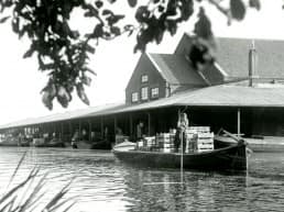 historisch zwart-wit foto van een tuinder die met een stok zijn platte boot wegduwt bij de veiling in het Westland met op zijn boot houten kisten