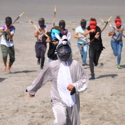 deelnemers aan de groepsactiviteit 'Shoot Out' van Wato Events rennen met pijl en boog aan achter iemand die verkleed is in een konijnenpak op het strand in het Westland