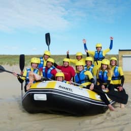 foto van een groep enthousiaste mensen die gaan raften in wetsuits en met gele helmen en roeispanen in een rubberen boot op het strand van Ter Heijde in het Westland