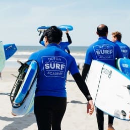 grosp sufers met zwarte wetsuits en blauwe t-shirt lopen op het strand met surfboards onder de arm richting de zee in het Westland